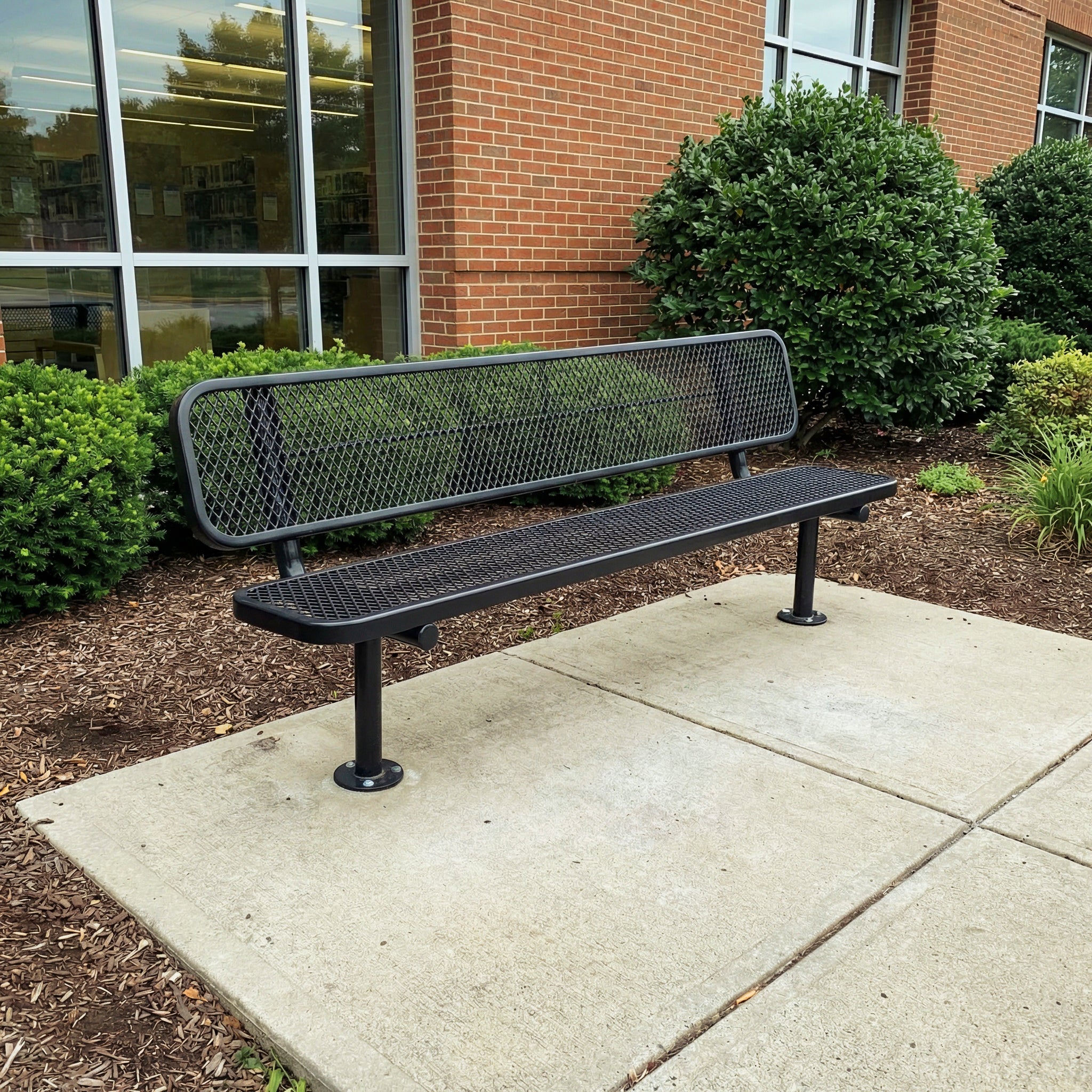 Black metal bench on a concrete pavement with a brick building and greenery in the background