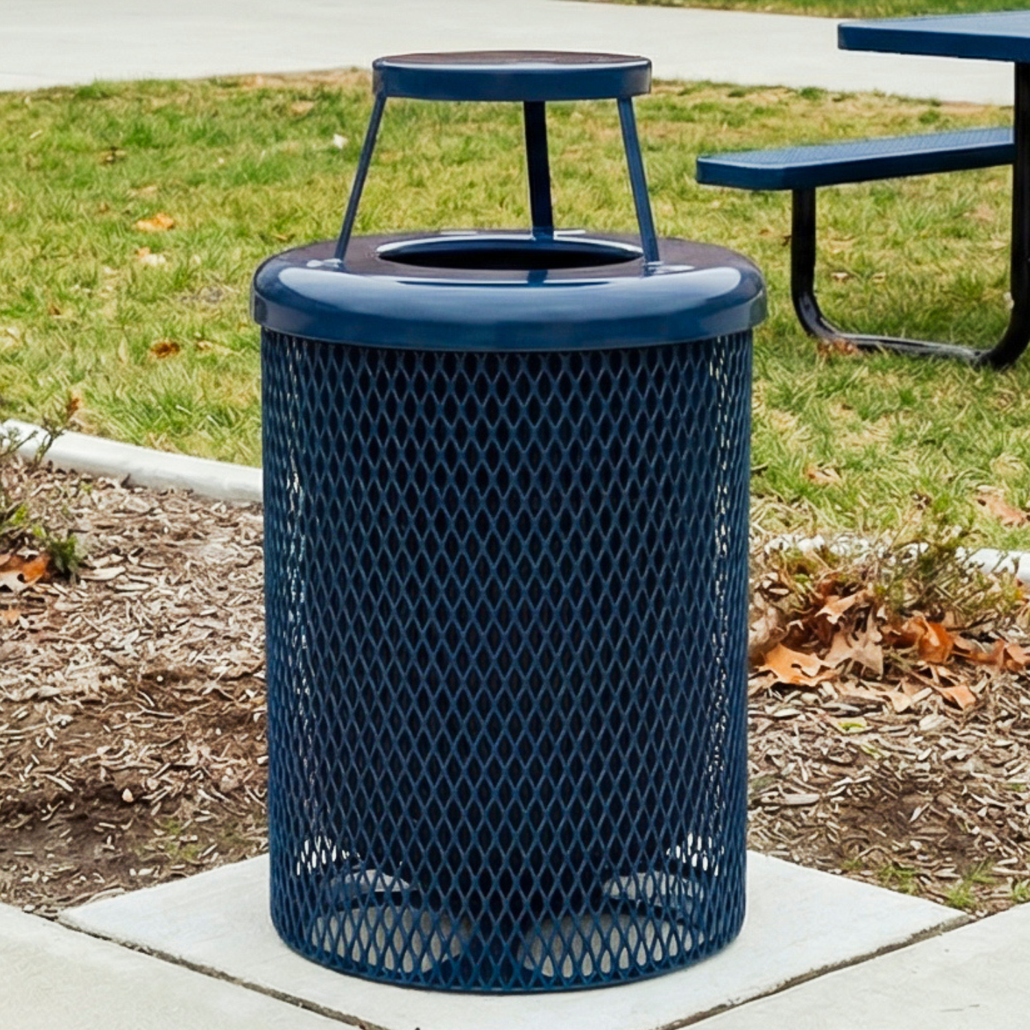 Blue mesh trash can on a sidewalk with grass and a picnic table in the background
