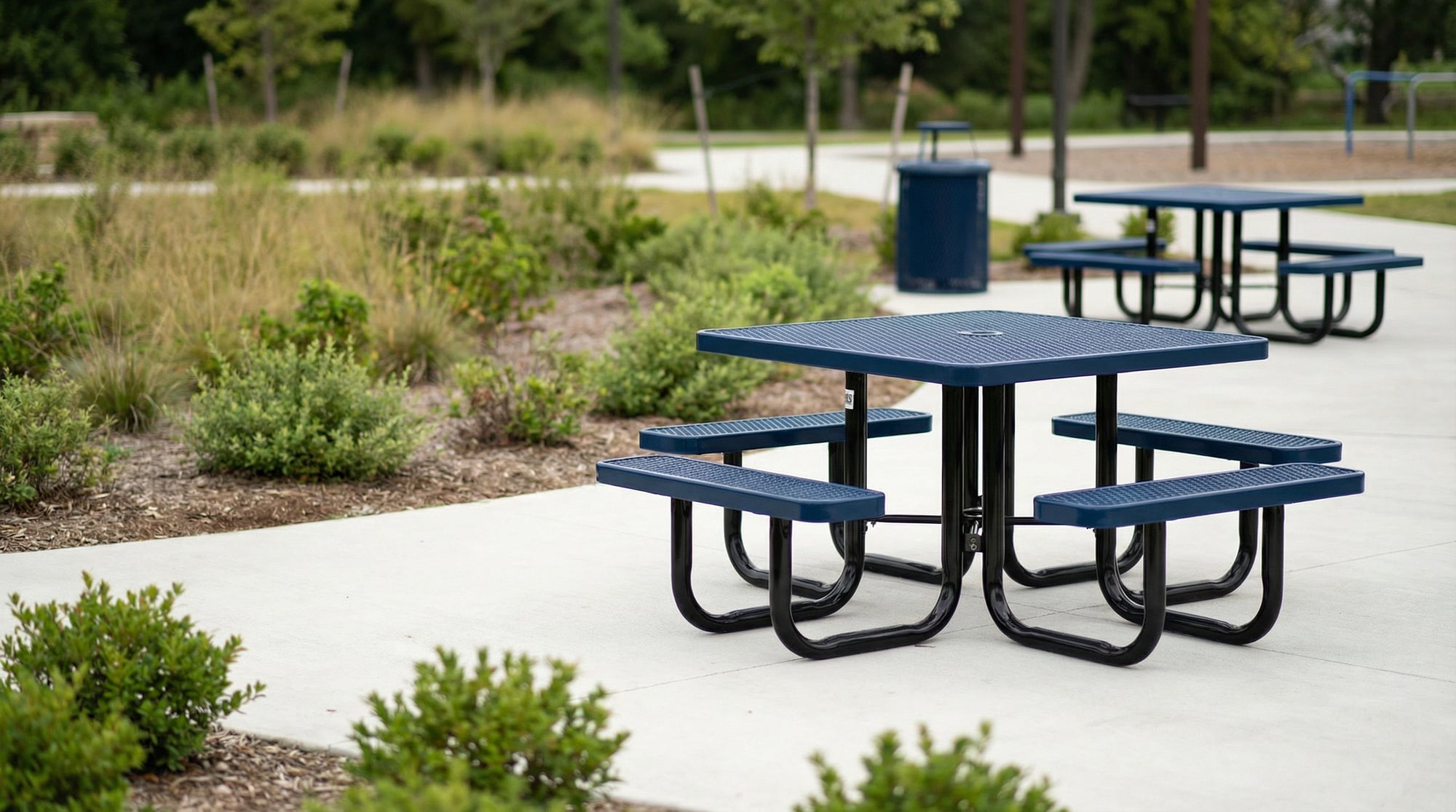 Dark blue heavy-duty expanded metal picnic table with matching trash receptacle installed in a public park setting