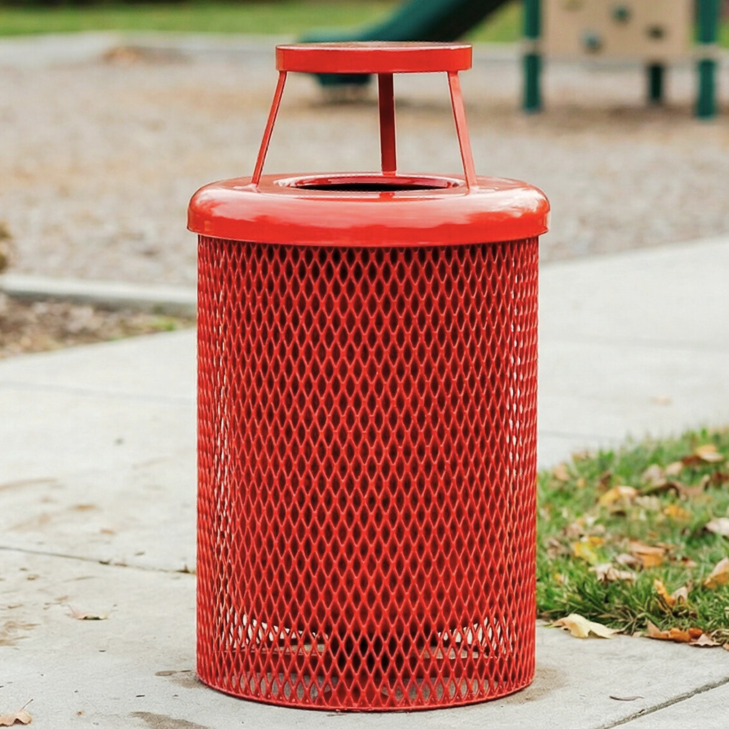 Red metal trash can with a textured surface on a paved area with grass and playground equipment in the background.