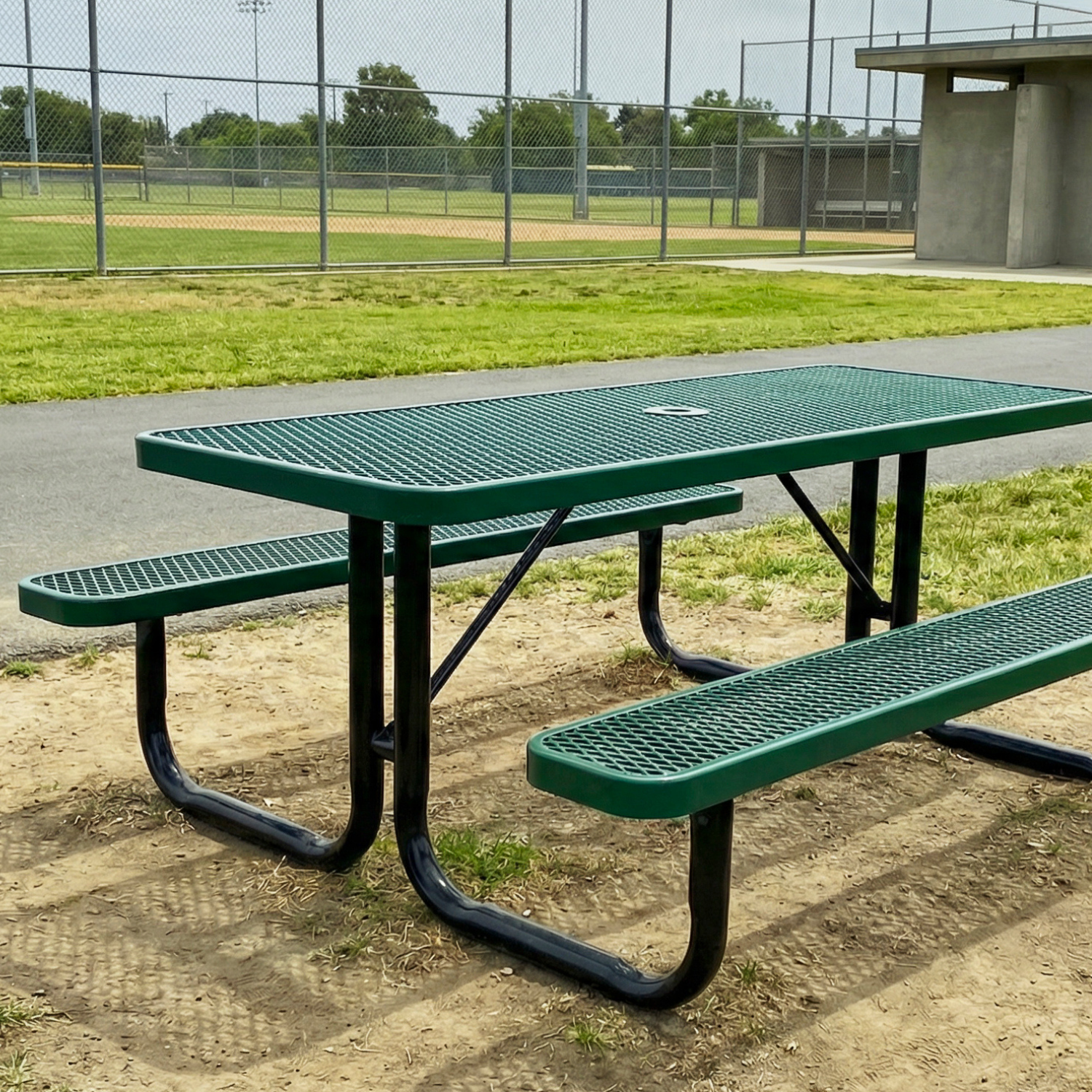 Green metal picnic table with benches on a dirt surface, with a baseball field in the background.