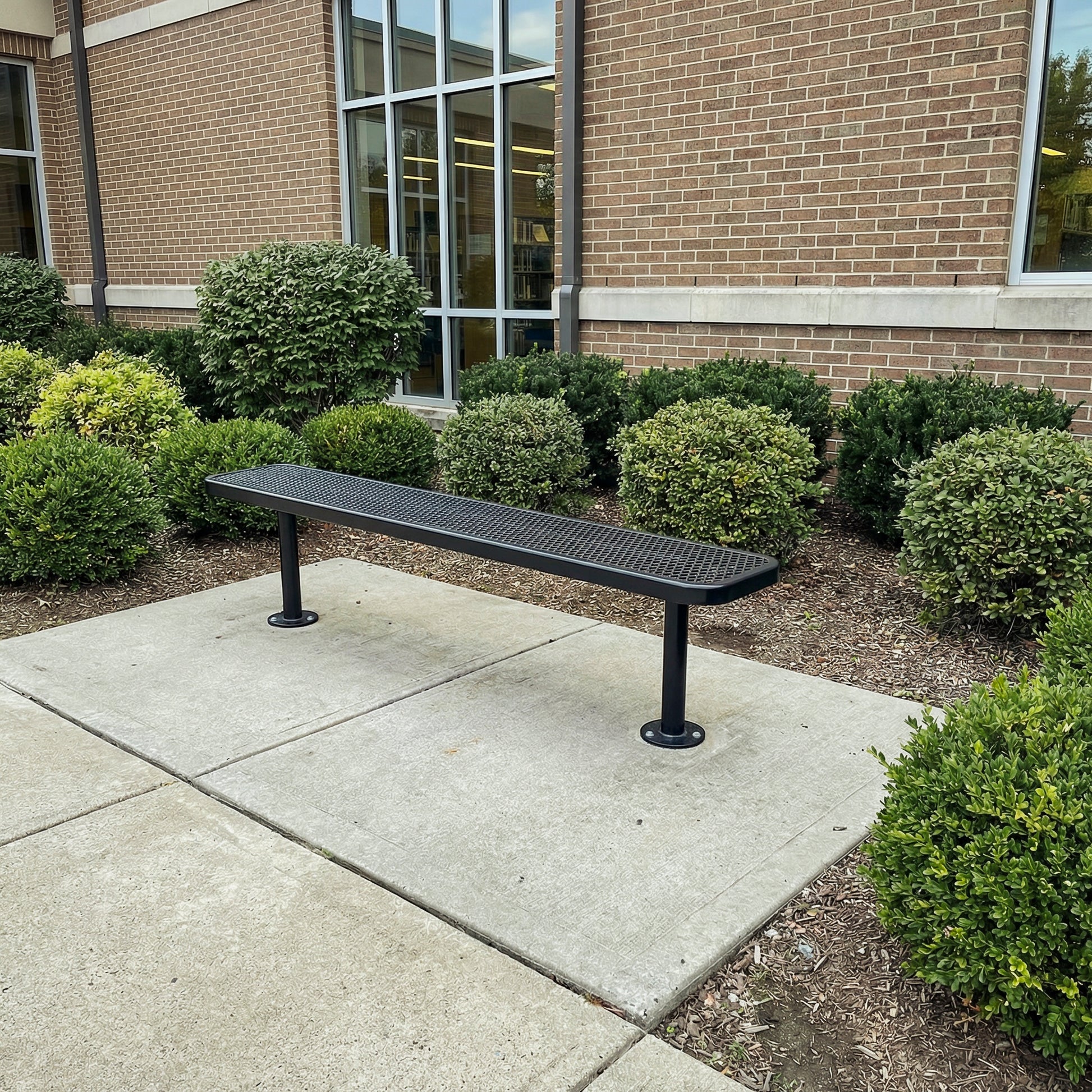 Black metal bench on a concrete pavement with a brick building and greenery in the background